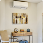 A modern room corner with a beige chair, a wooden console table displaying decorative vases and books, abstract wall art, and a sleek white mini split installation mounted above.