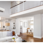 Modern, airy living room with a white sofa, patterned pillows, two tan lamps, wooden floors, and open view to a bright kitchen and upstairs balcony railing. A wall-mounted mini split installation is visible above the main area.