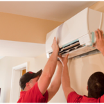 Two workers in red shirts and black caps are performing a mini split installation, servicing a wall-mounted air conditioning unit inside a beige room with natural light streaming through a window.