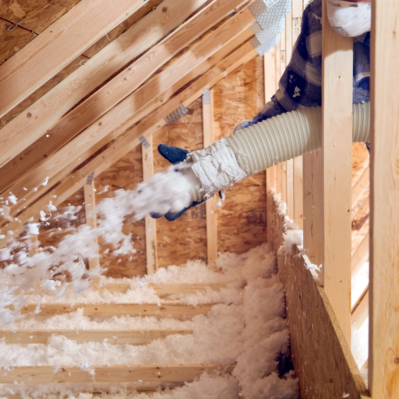 A person uses a large hose to blow loose-fill insulation material into an unfinished attic with exposed wooden beams. Spray Foam Insulation Installers often offer similar services to ensure fluffy insulation covers every inch of the floor.