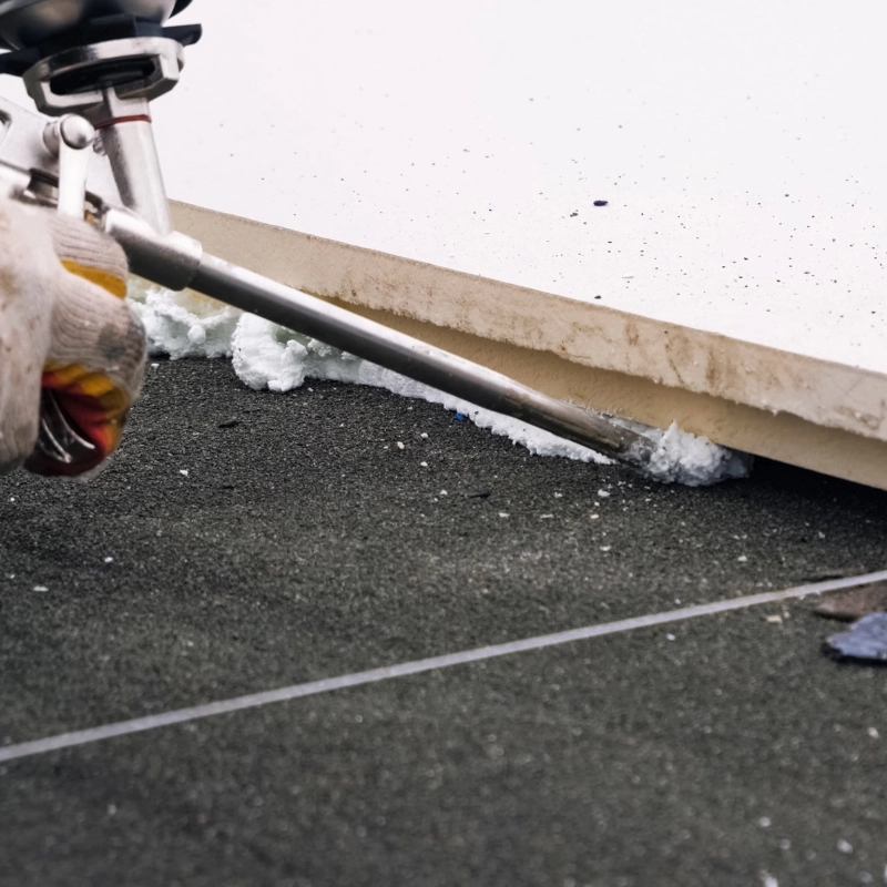 A gloved hand uses a caulking gun to apply foam insulation along the base of a wall on a dark, textured surface, showcasing the precision of spray foam insulation installers.