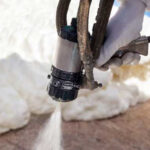 A gloved hand sprays expanding foam insulation onto a surface using a spray gun, showcasing the expertise of Spray Foam Insulation Installers with fresh white foam visible in the background.