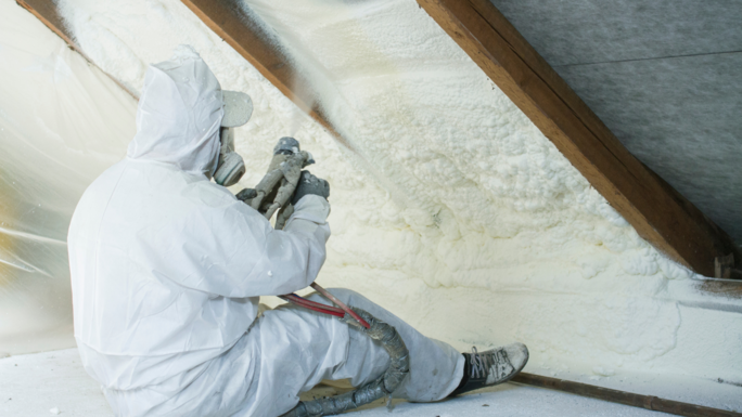Builder sitting in the attic, spraying the roof above him with foam to seal it