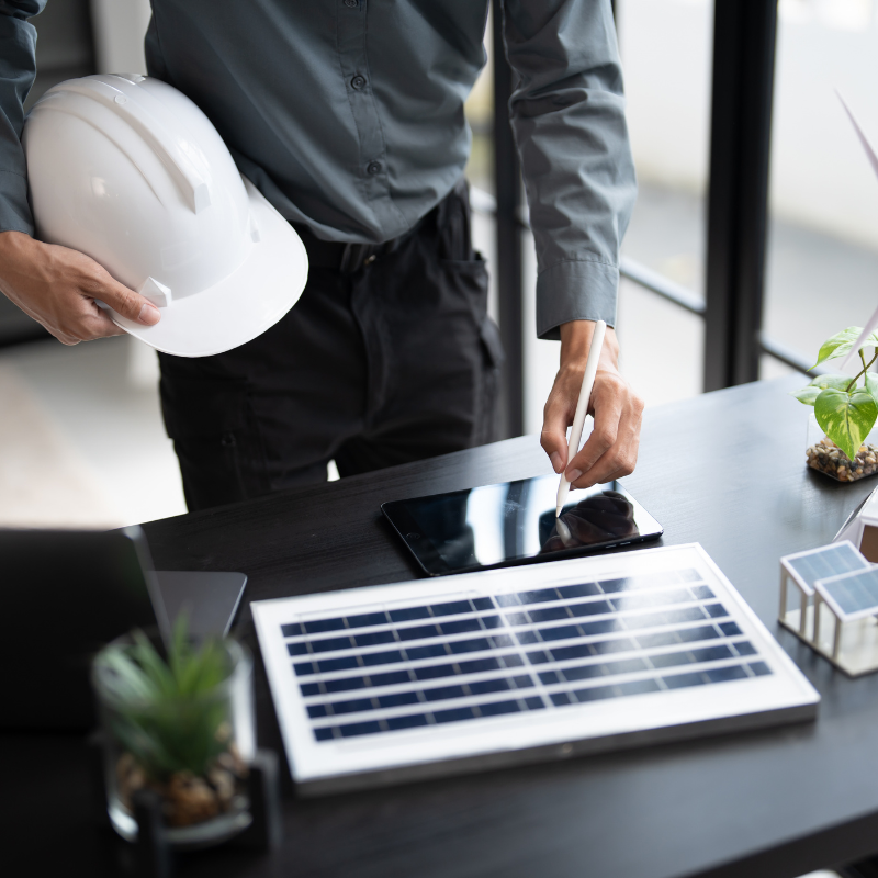 A person standing at a desk holds a white hard hat and uses a tablet. On the desk are a small solar panel, laptop, miniature house, and potted plants, suggesting involvement in renewable energy or Mass Save Program projects.
